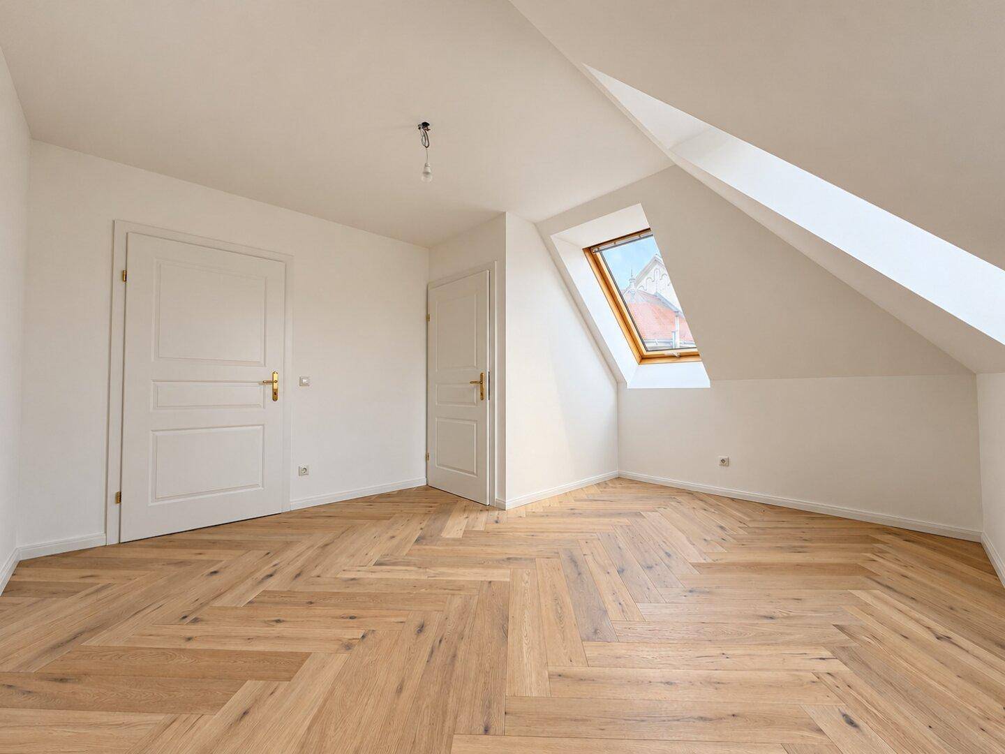Unmöblierter Raum mit Fischgrätparkett, Dachschräge und einem Dachfenster für natürliches Licht.
