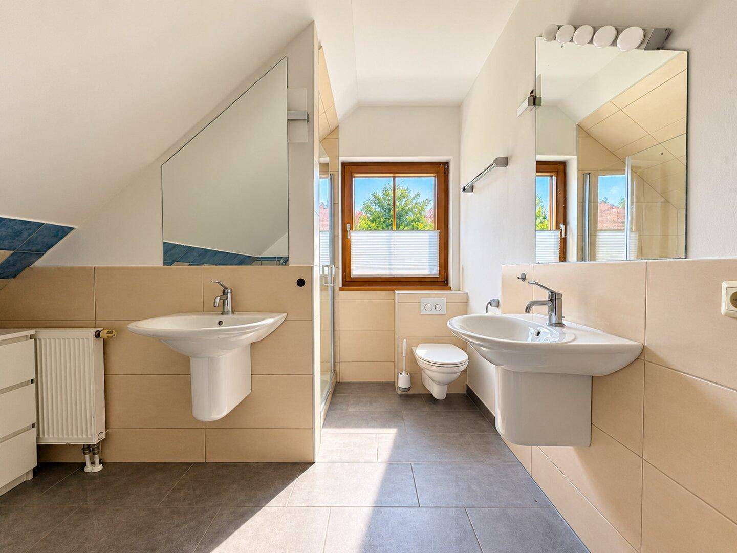 Geräumiges Badezimmer mit zwei Waschbecken, Toilette und Fenster für Tageslicht.