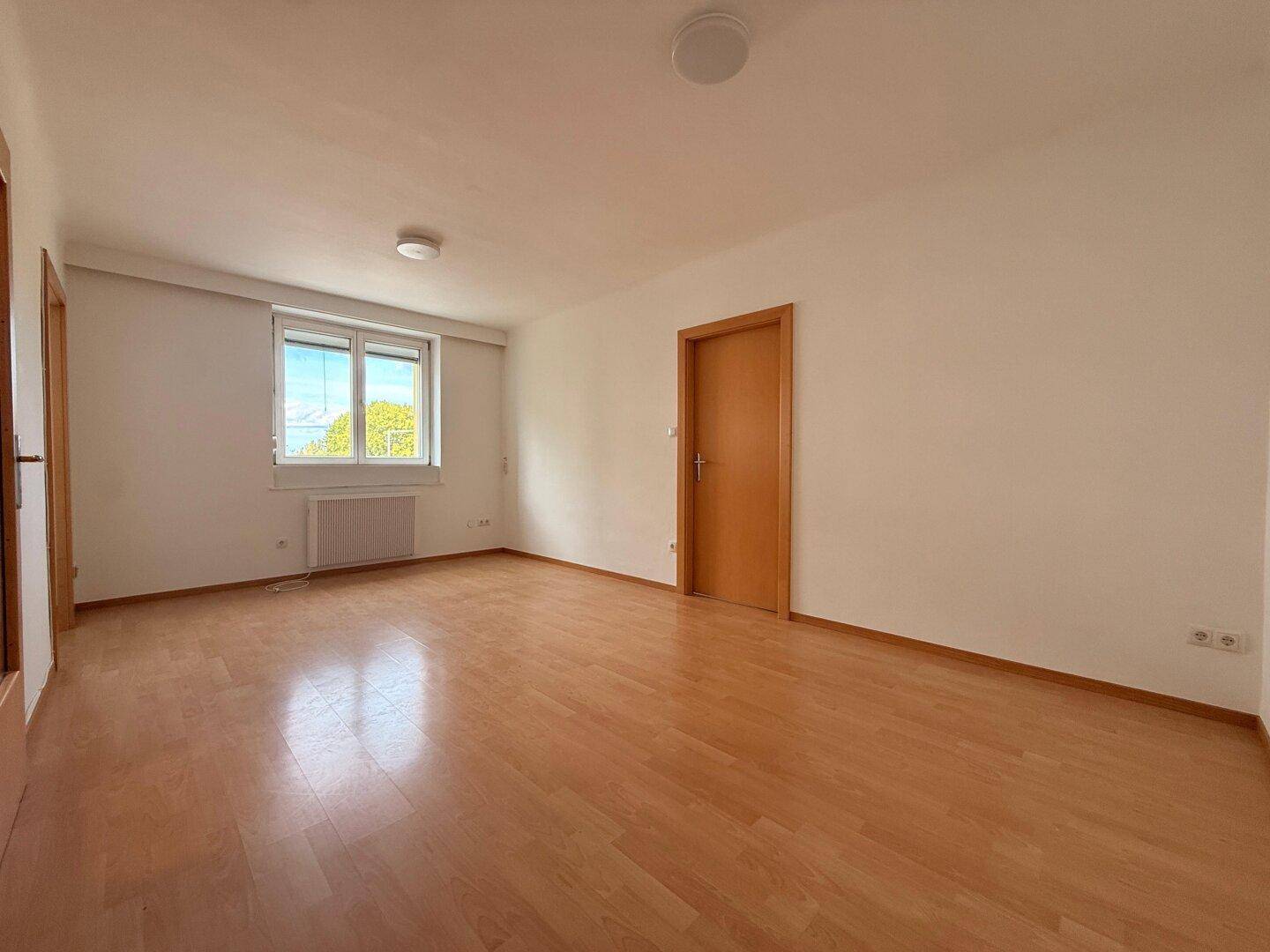 Geräumiges, leeres Wohnzimmer mit hellem Parkettboden und großem Fenster.