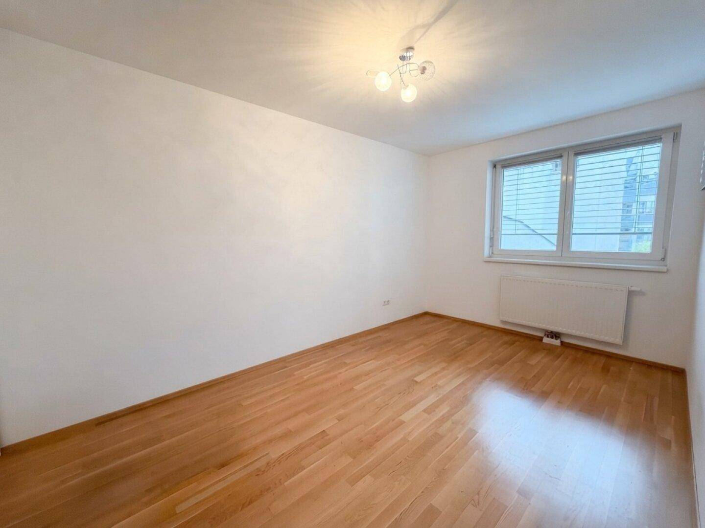 Helles, leeres Zimmer mit Parkettboden und großem Fenster, ideal als Schlaf- oder Arbeitszimmer.