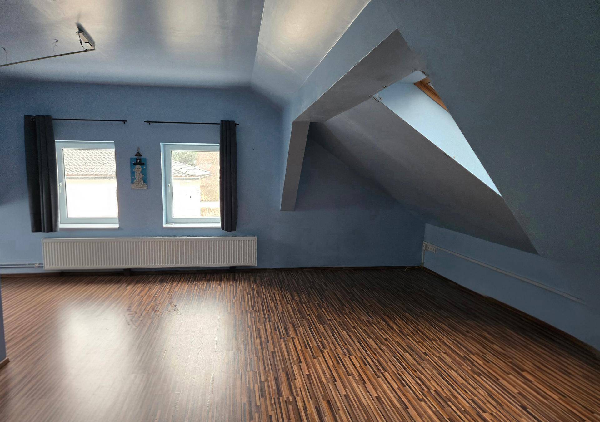 Geräumiges Dachzimmer mit blauen Wänden, Holzboden und zwei Fenstern.