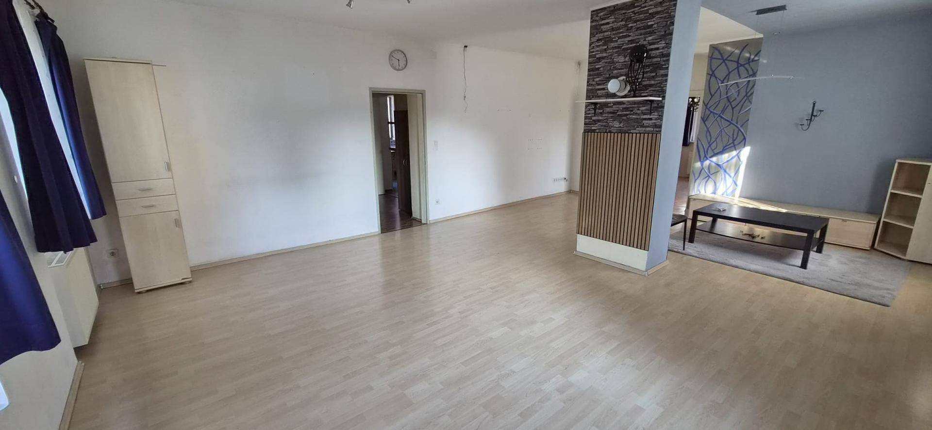 Geräumiges, leeres Wohnzimmer mit hellem Laminatboden und strukturiertem Wandelement.