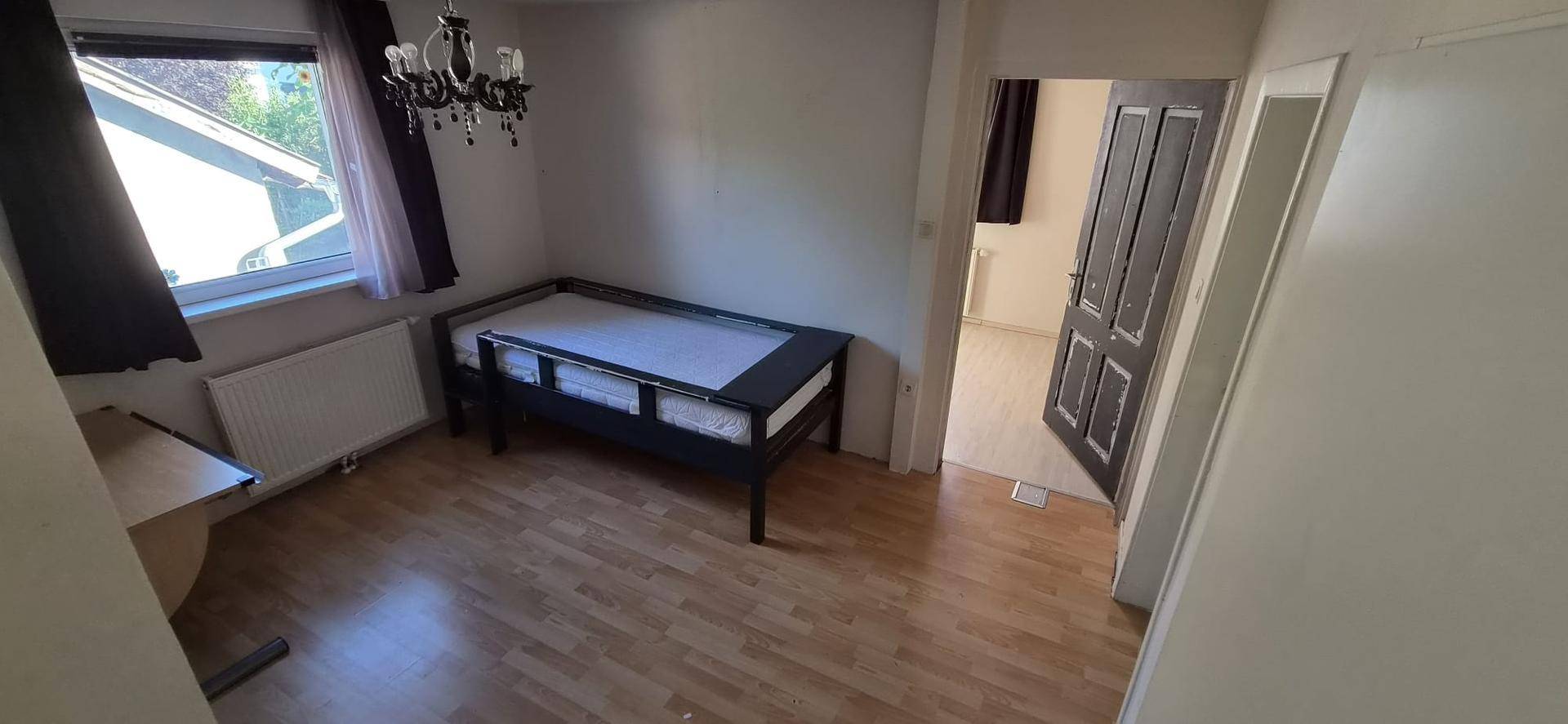 Helles Zimmer mit Holzboden, Fenster und einfacher Bettkonstruktion.