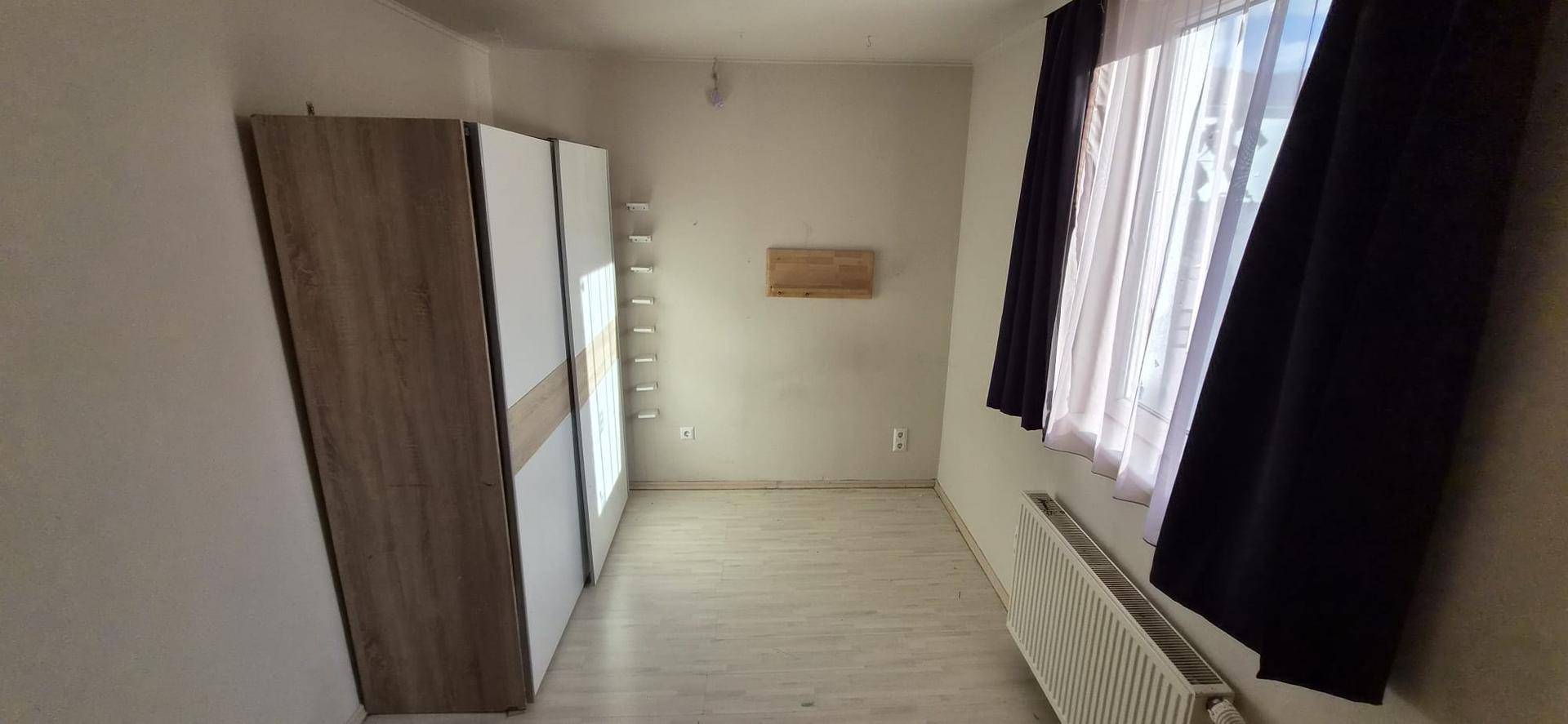 Leeres Zimmer mit hellem Boden, großem Fenster und Heizkörper.