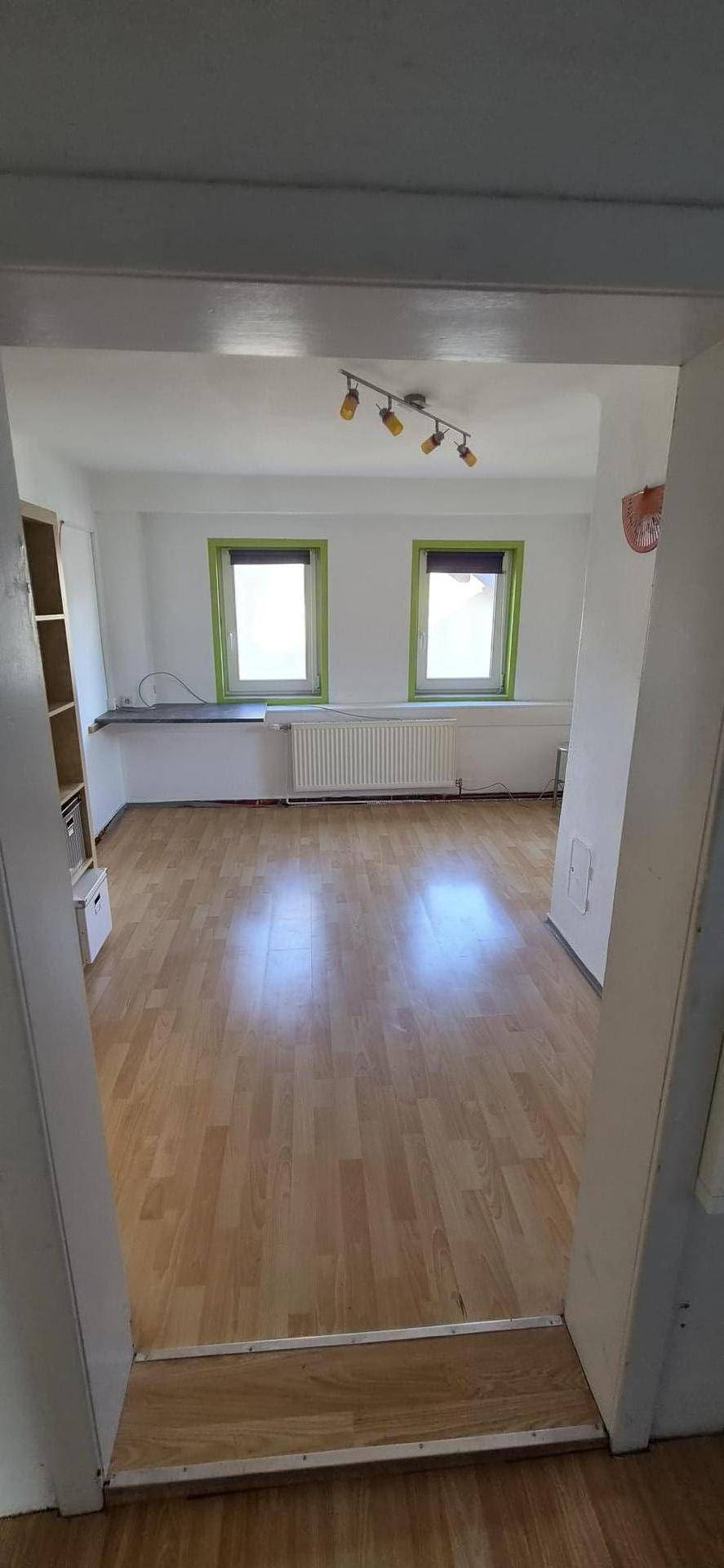 Helles Zimmer mit Holzboden und zwei Fenstern mit grünen Rahmen.