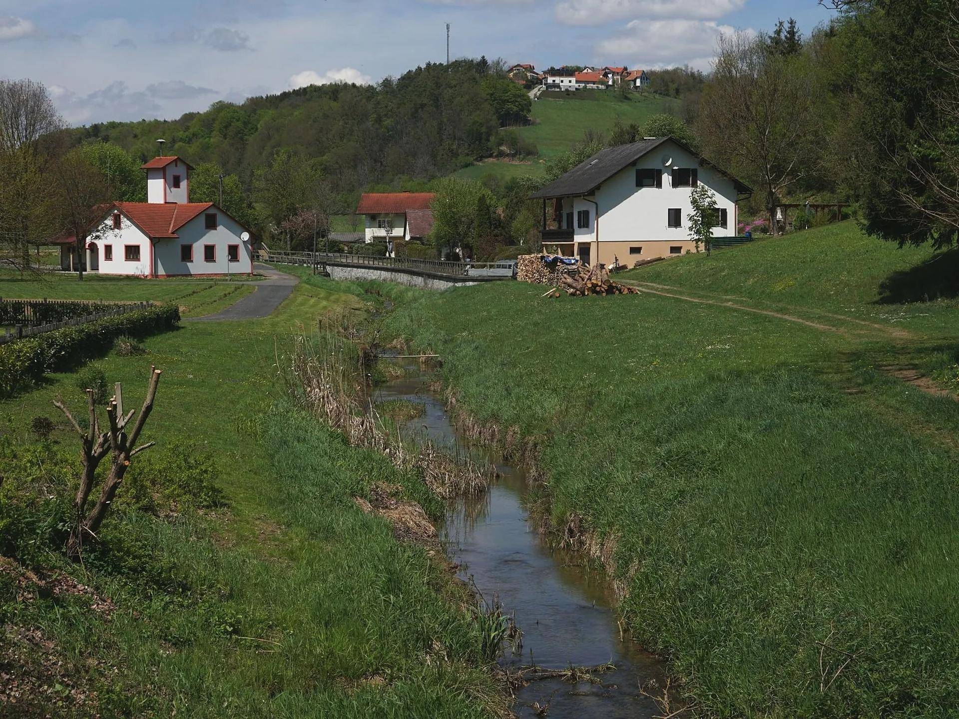 Idyllische Landschaft mit einem kleinen Bach, grünen Wiesen und Häusern am Hügel.