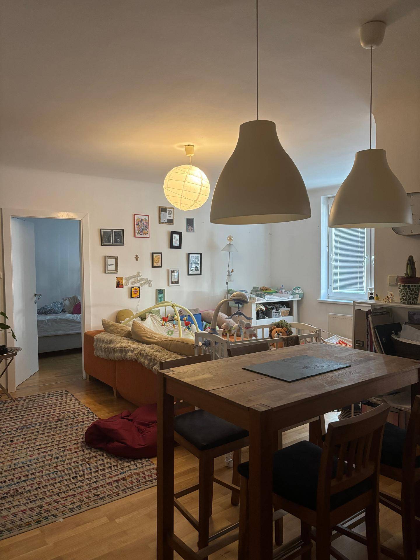 Heller Wohn-Essbereich mit Holztisch, Hängelampen und einem Babybett im Hintergrund.