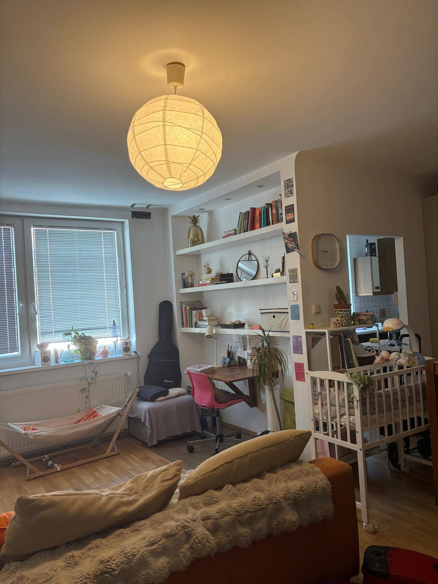 Heller Wohnbereich mit Schreibtisch, Bücherregal und einem Babybett am Fenster.