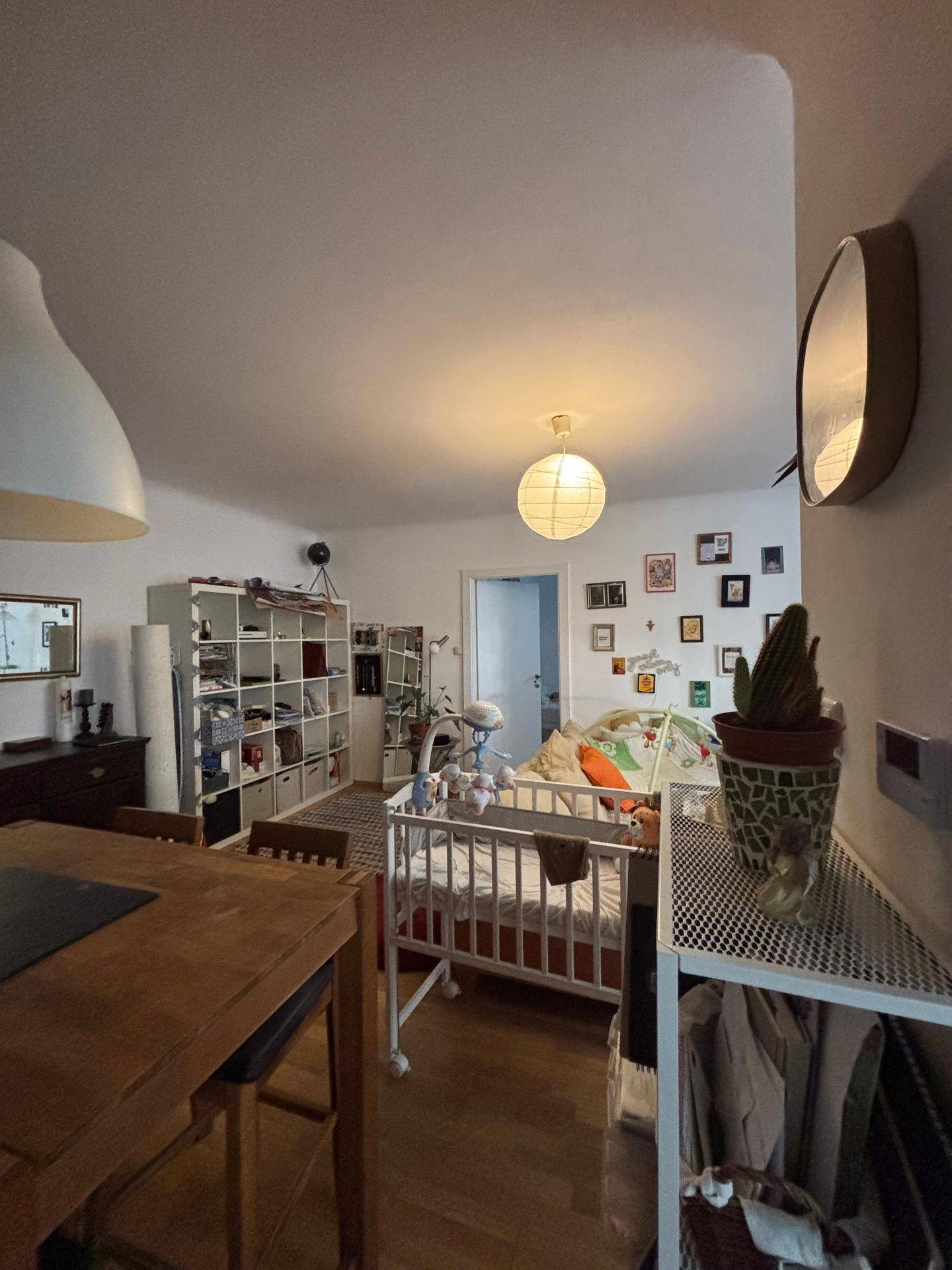 Geräumiger Wohn-Essbereich mit Holzboden, großem Regal und Babybett im Vordergrund.
