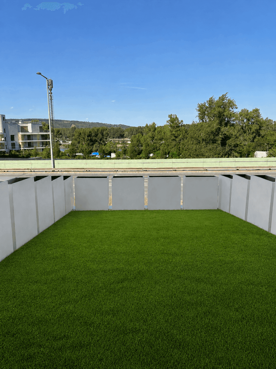 Kleiner, ummauerter Gartenbereich mit Kunstrasen und weitem Blick auf grüne Bäume und Gebäude unter blauem Himmel.