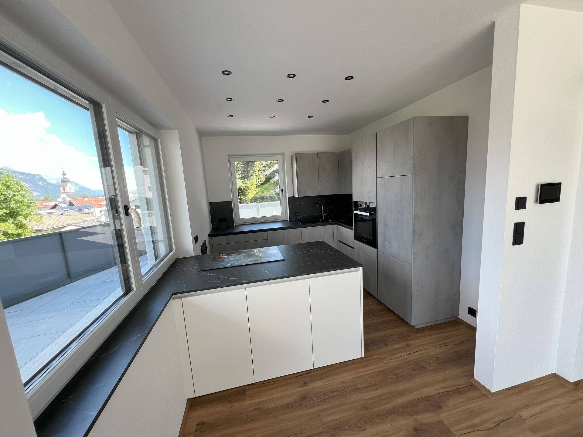 Moderne Küche mit grauen und weißen Schränken, Kochinsel und großen Fenstern mit Ausblick.