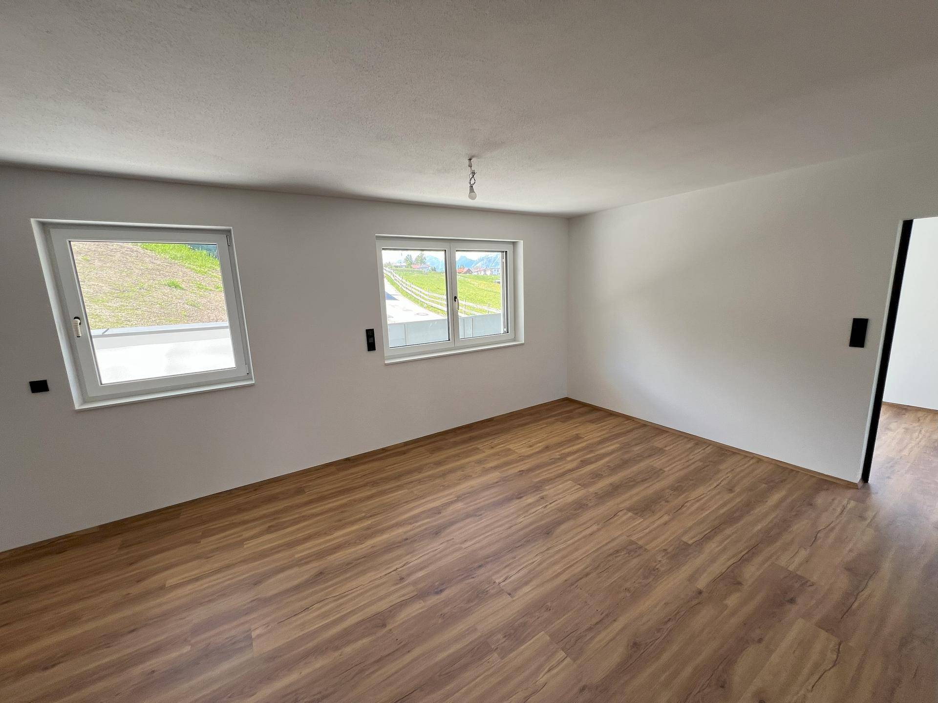 Leerer Raum mit Laminatboden, weißen Wänden und zwei Fenstern, die viel Licht hereinlassen.