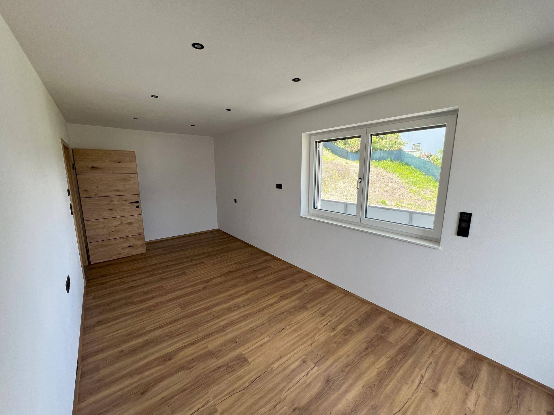 Leerer Raum mit Laminatboden, weißen Wänden und einem Fenster mit Blick ins Grüne.