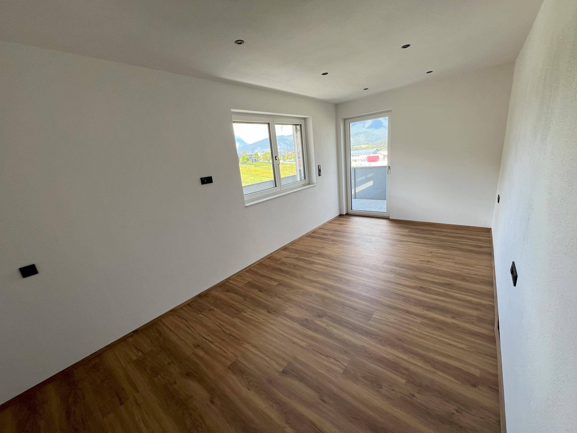 Heller Raum mit Laminatboden, weißen Wänden und Zugang zu einem Balkon oder einer Terrasse.