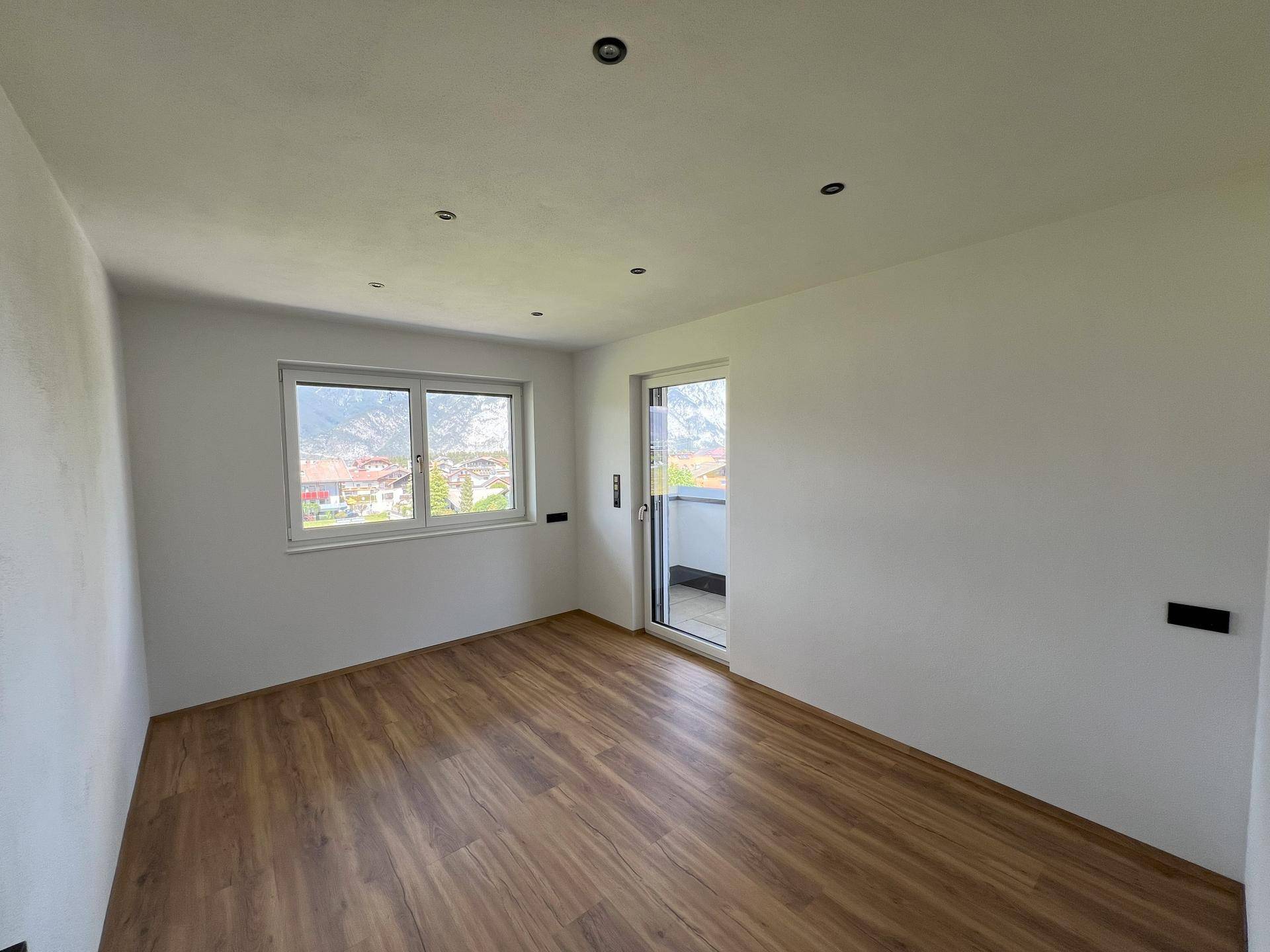Geräumiger Raum mit Laminatboden, weißen Wänden und Zugang zu einem Balkon oder einer Terrasse.