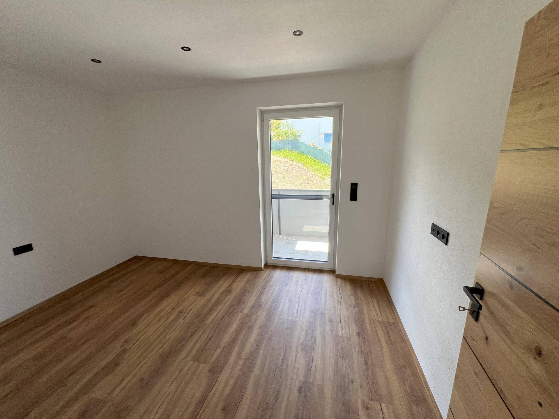 Heller Raum mit Laminatboden und Zugang zu einem Balkon oder einer kleinen Terrasse.