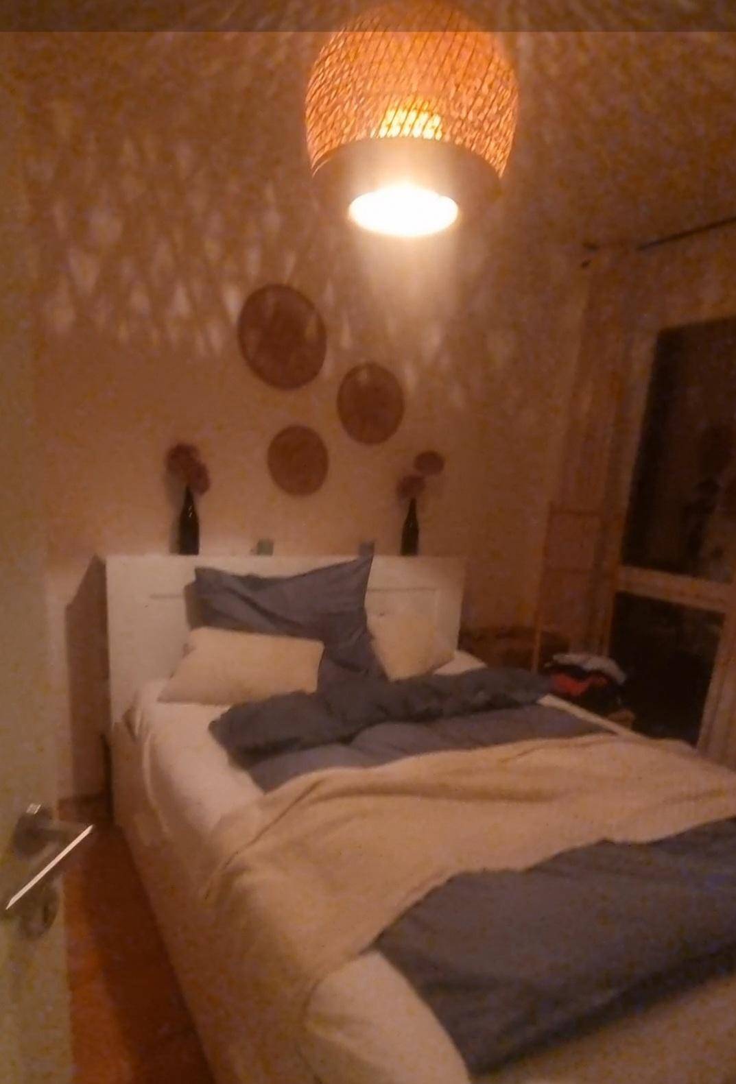 Schlafzimmer mit Doppelbett, Holzboden und einer dekorativen Korblampe, die Muster an die Wand wirft.