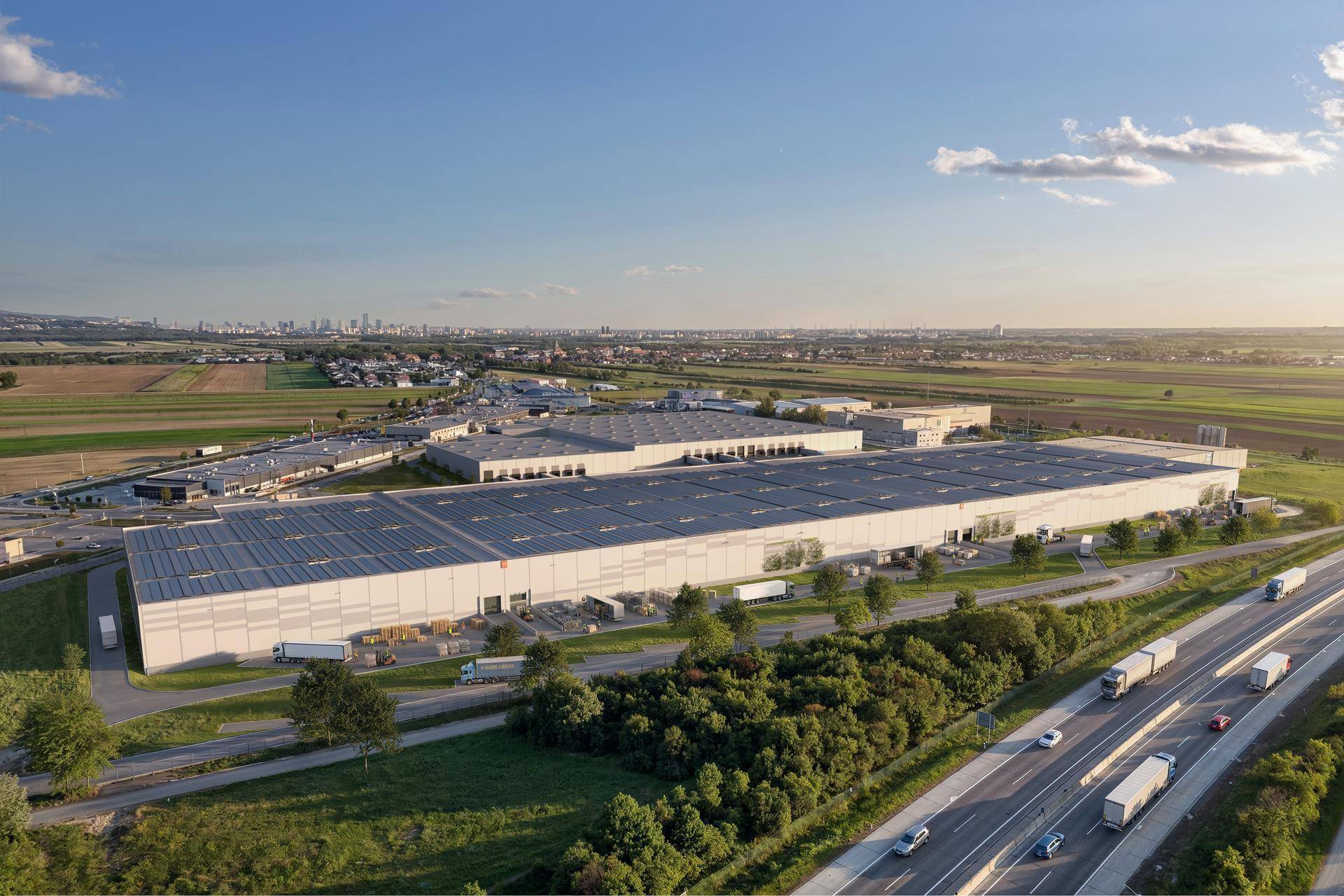 Luftaufnahme des großen Logistikparks mit Solaranlagen auf den Dächern, nahe einer Autobahn und Stadt.