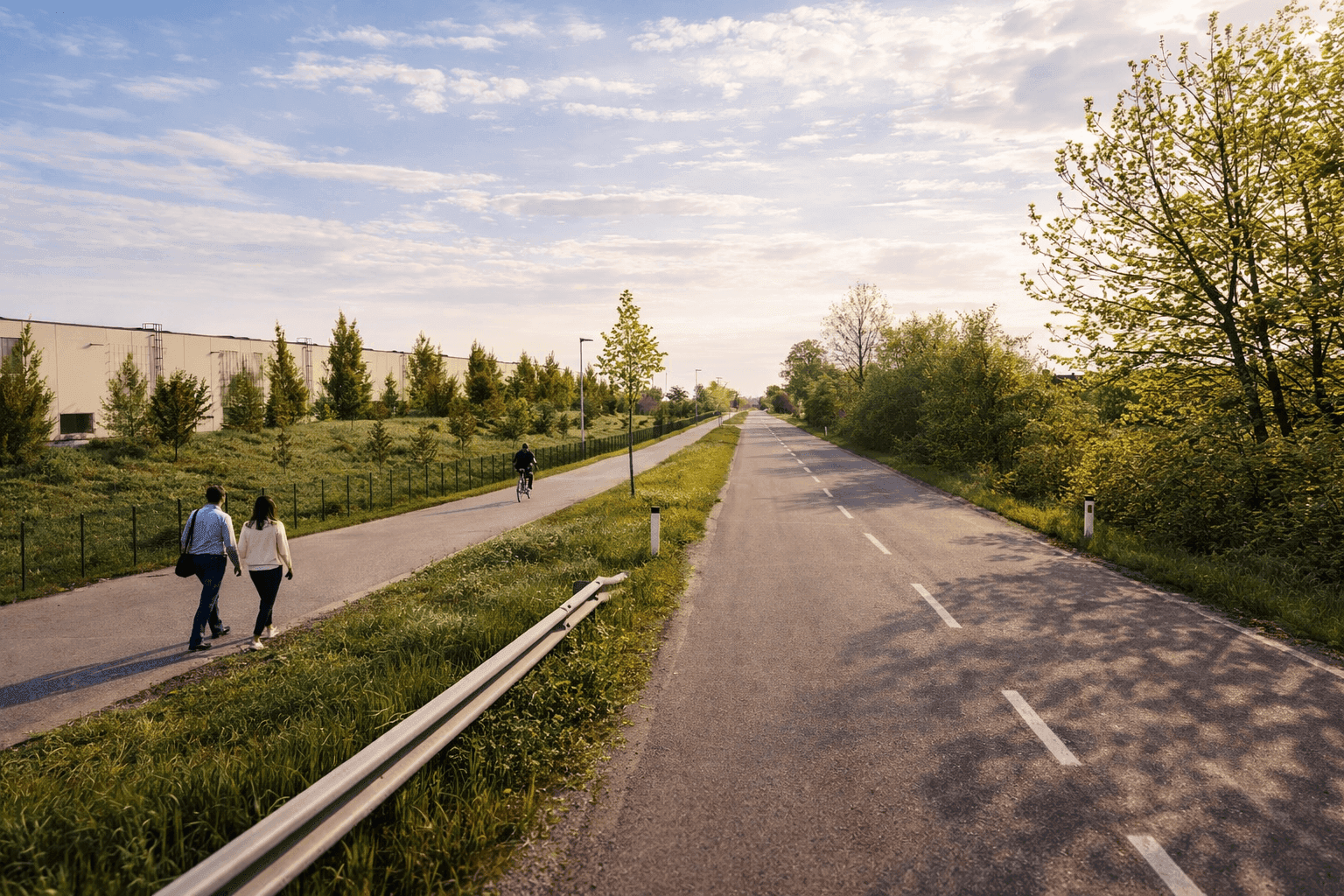 Lange Straße mit separatem Rad- und Fußweg, umgeben von grünen Feldern und Bäumen. Ein großes Gebäude ist in der Ferne sichtbar.