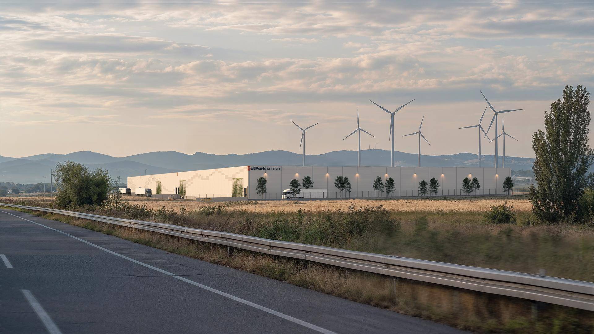 Weitläufige Ansicht des Logistikzentrums von der Straße, umgeben von Feldern und Windrädern.