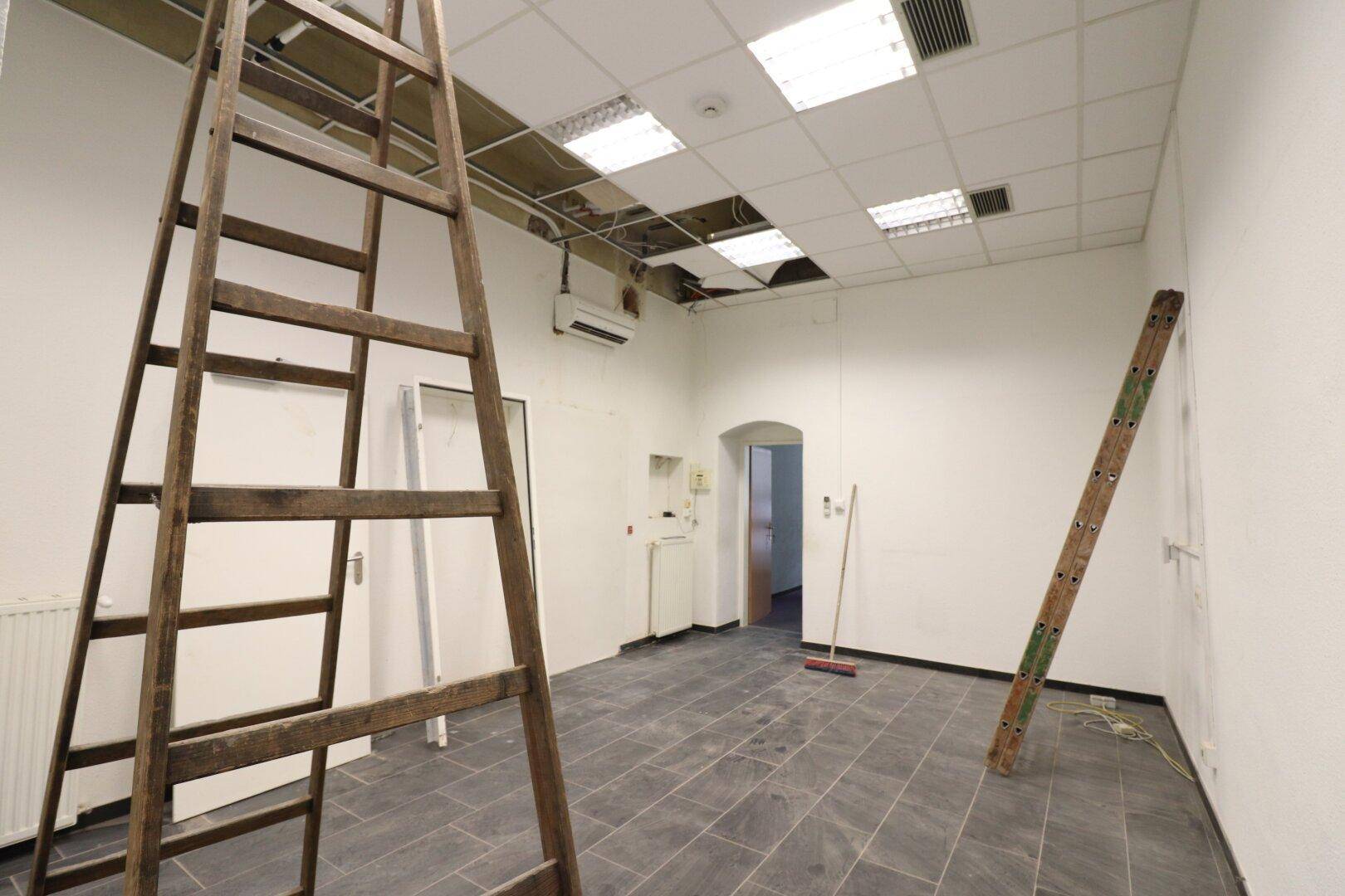 Leerer Gewerberaum mit Fliesenboden, abgehängter Decke und sichtbaren Leitungen während der Renovierung.
