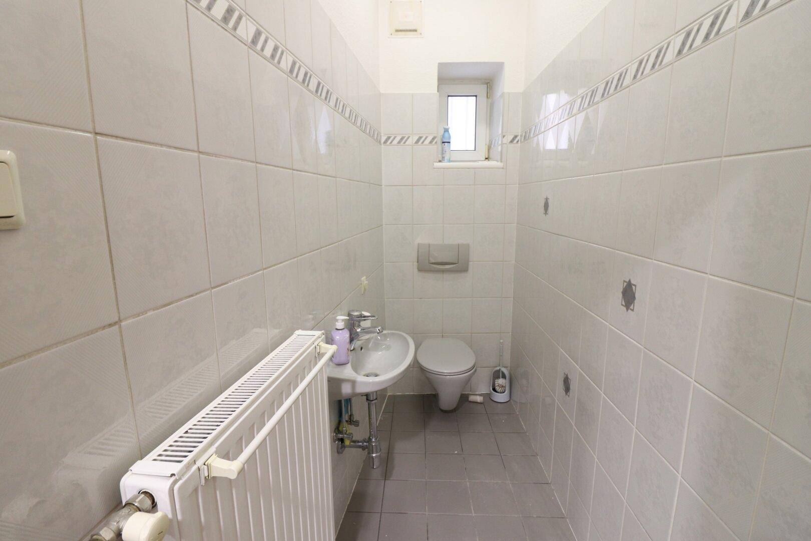 Kleines, gefliestes Gäste-WC mit wandhängender Toilette, Waschbecken, Heizkörper und Fenster.
