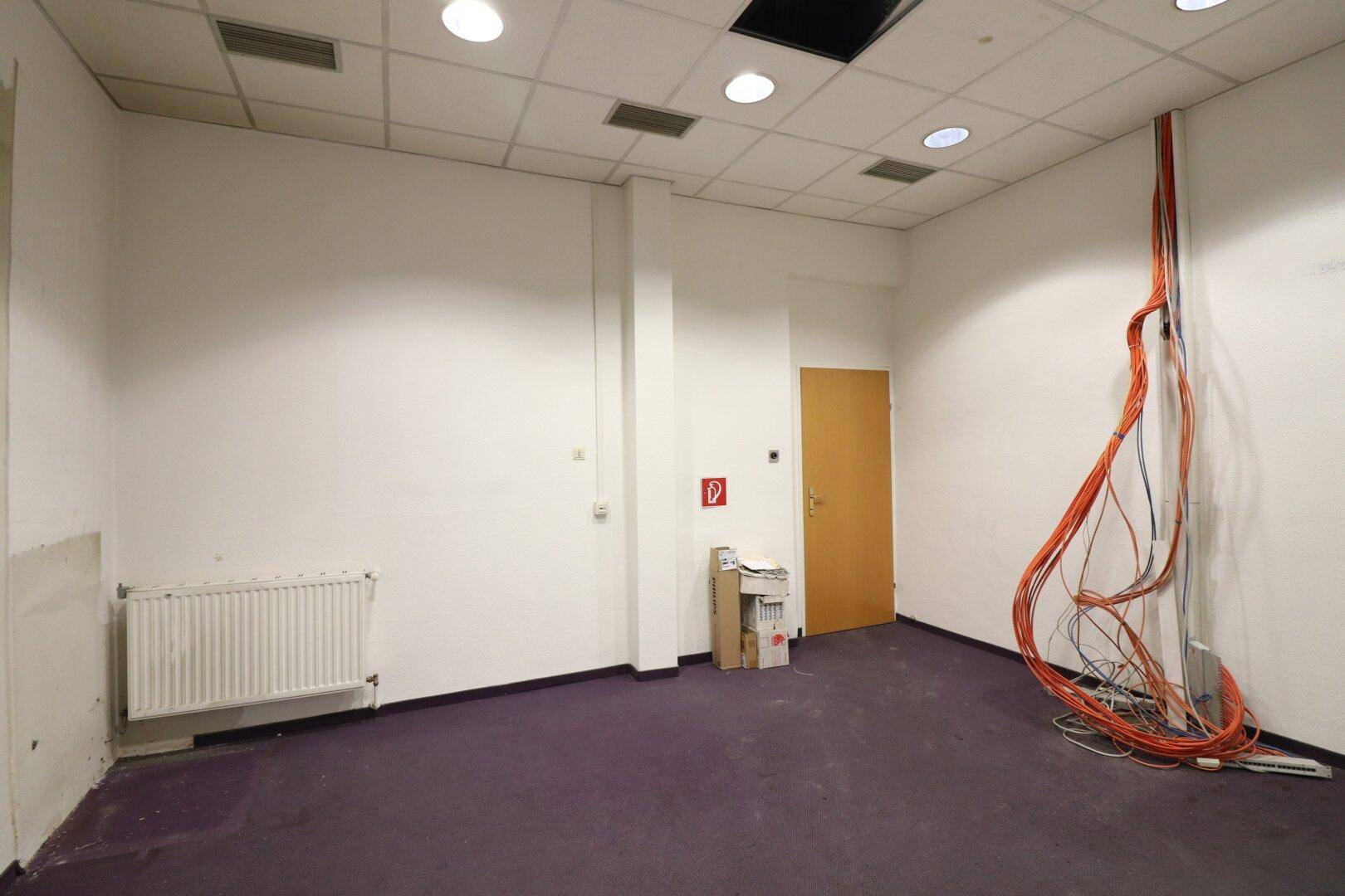 Leerer Gewerberaum mit lila Teppichboden, Heizkörper und sichtbaren Kabeln an der Wand.