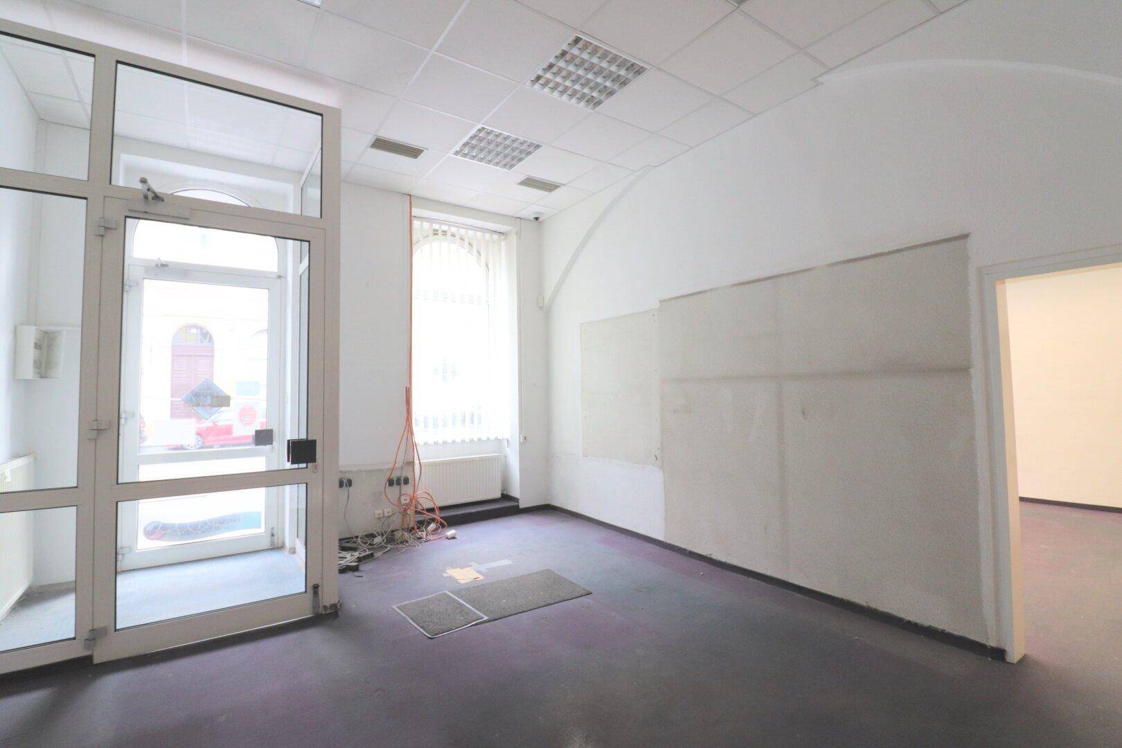 Leerer Gewerberaum mit großem Glaseingang, lila Teppichboden und Heizkörper unter dem Fenster.