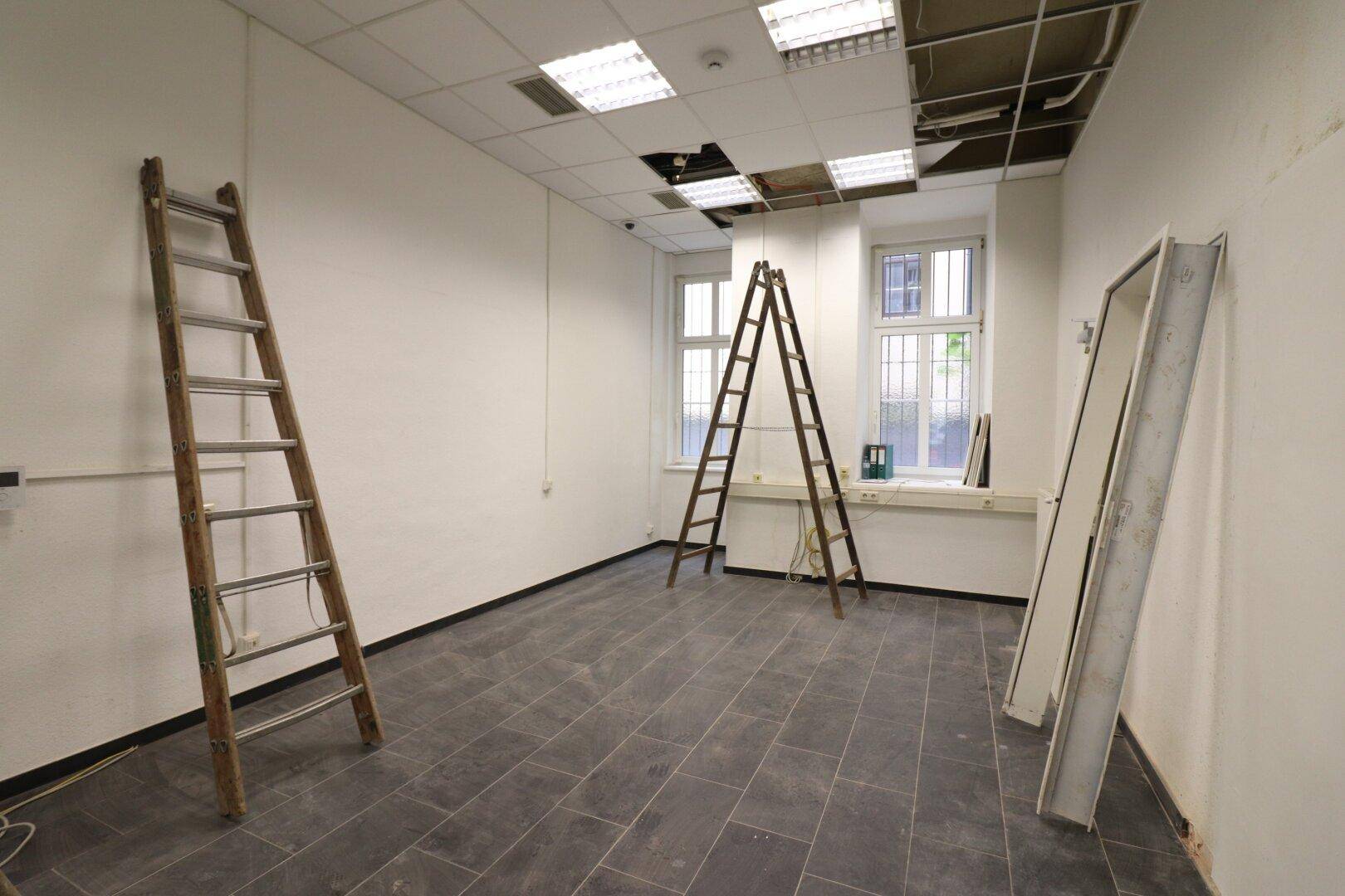 Leerer Gewerberaum mit Fliesenboden, zwei Leitern und Fenstern mit Gitterstäben während der Renovierung.
