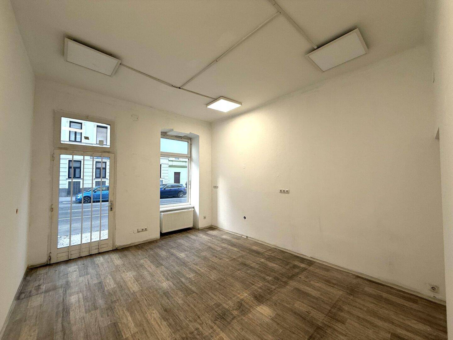 Heller Ladenraum mit Holzboden, weißen Wänden, großem Fenster und Glastür zur Straße.
