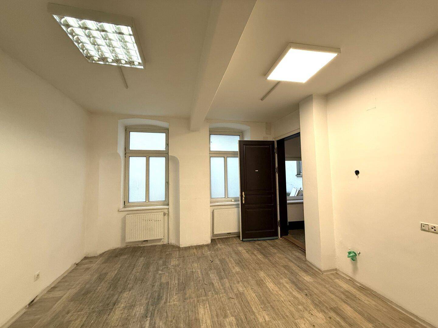 Leerer Büroraum mit Holzboden, weißen Wänden, zwei Fenstern und Deckenbeleuchtung.