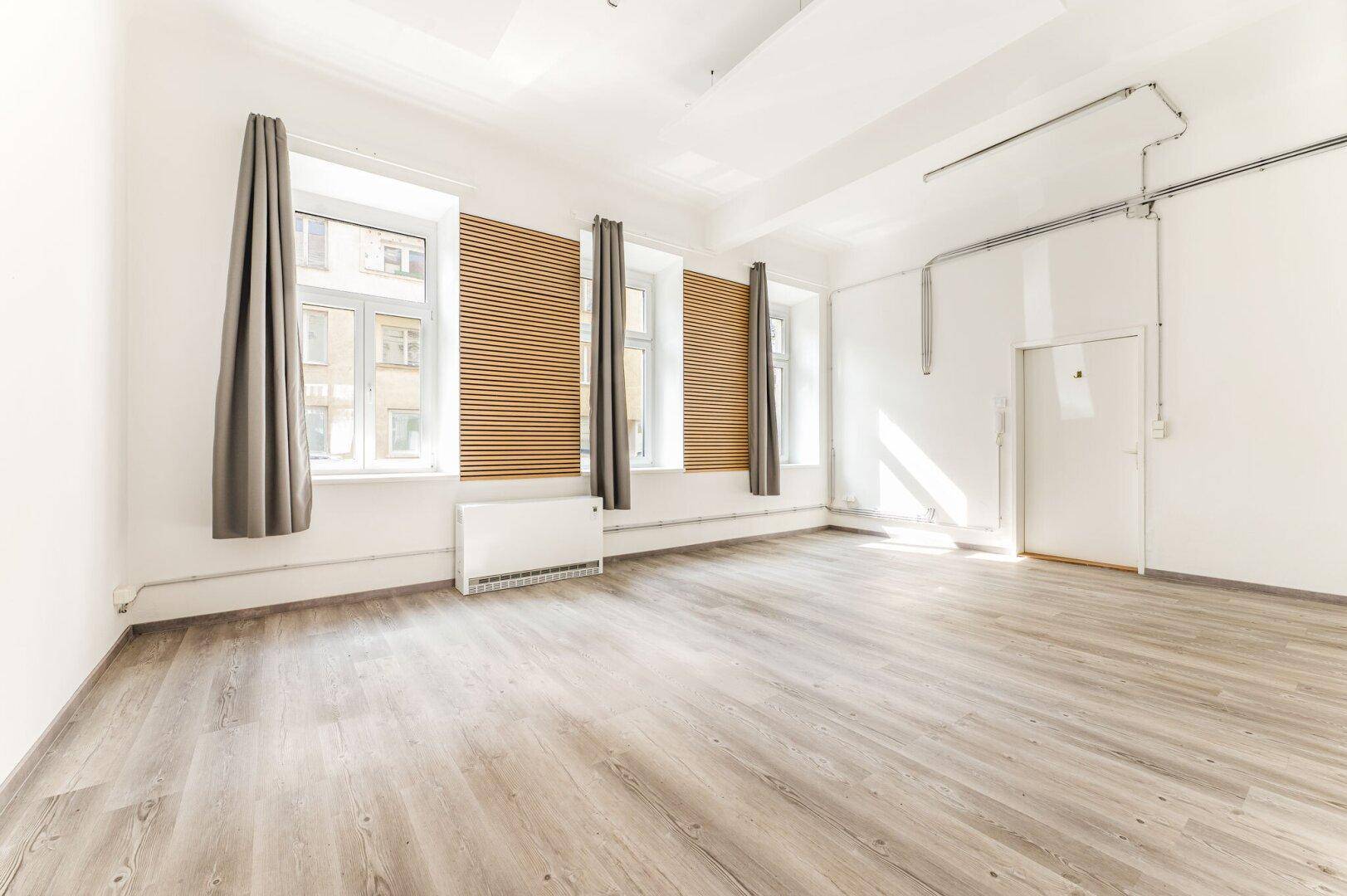 Heller, leerer Raum mit hohen Decken, großen Fenstern und modernem Laminatboden.