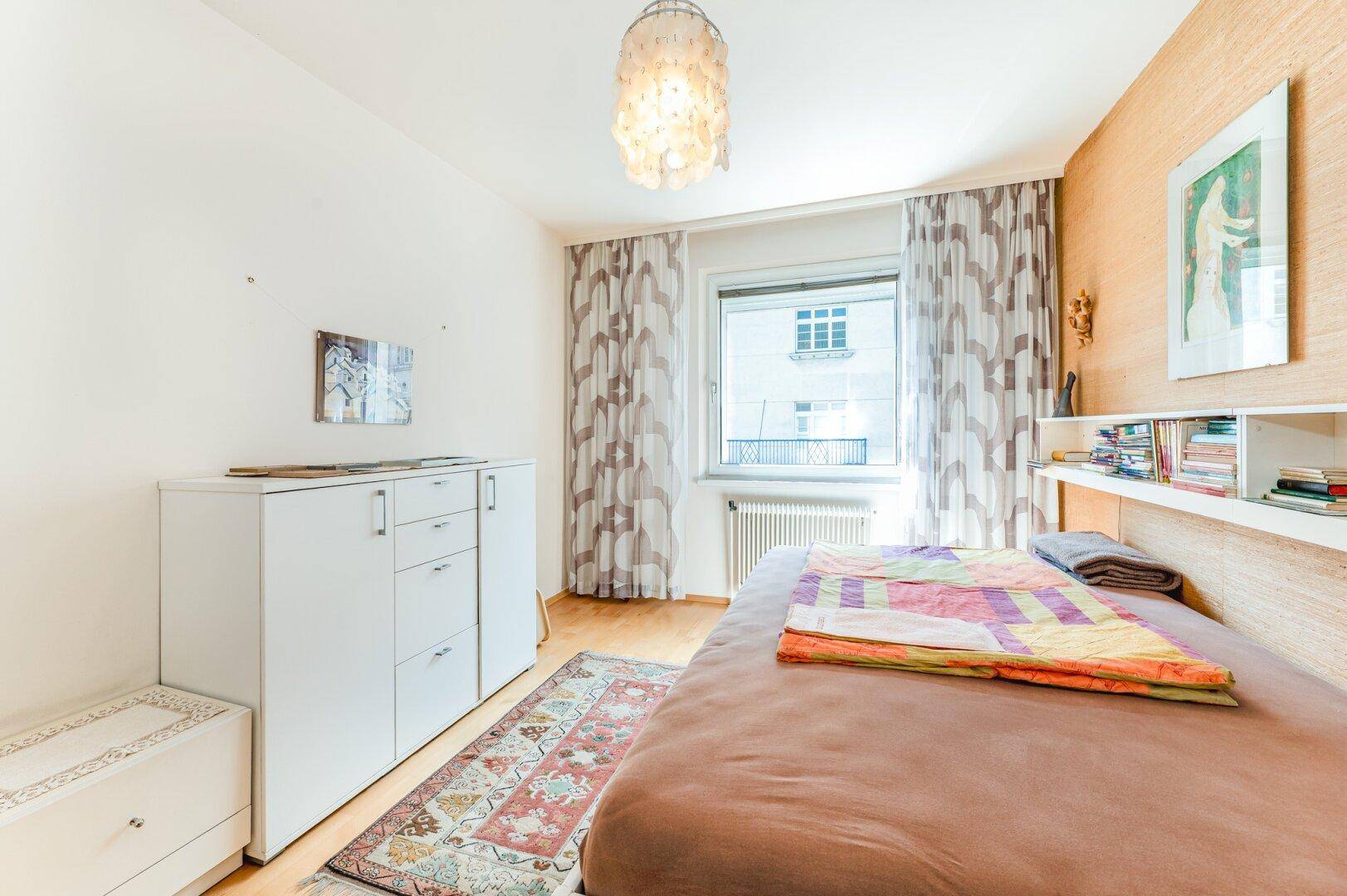 Schlafzimmer mit Holzboden, einem Bett, weißem Schrank und einem Fenster mit gemusterten Vorhängen.