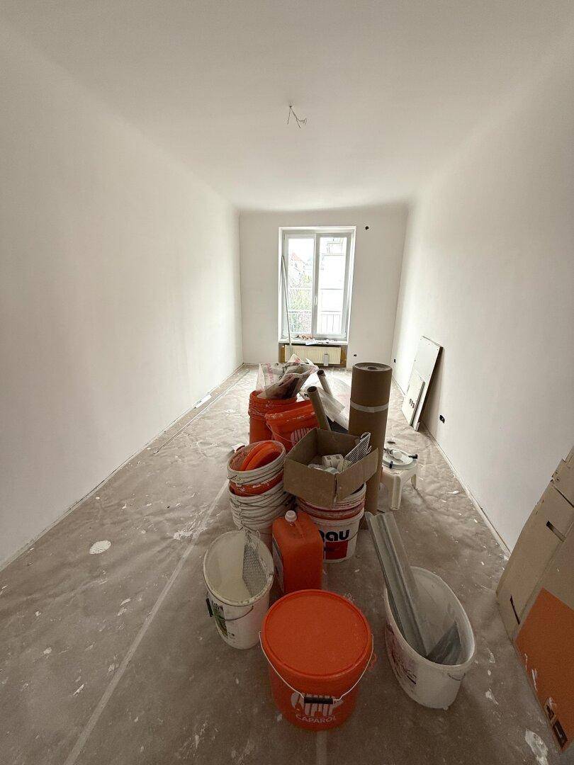 Renovierungsbedürftiger Raum mit weißen Wänden, einem Fenster und Baumaterialien auf dem abgedeckten Boden.
