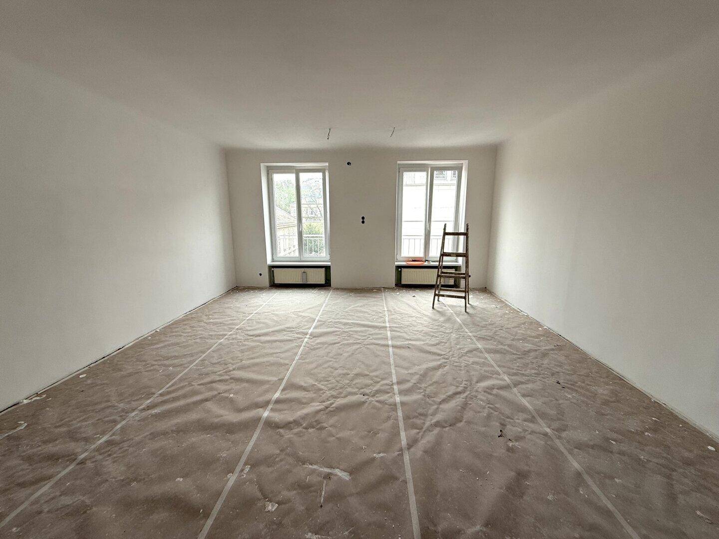 Geräumiger Raum in Renovierung mit zwei Fenstern, weißen Wänden und abgedecktem Boden, bereit für neue Einrichtung.