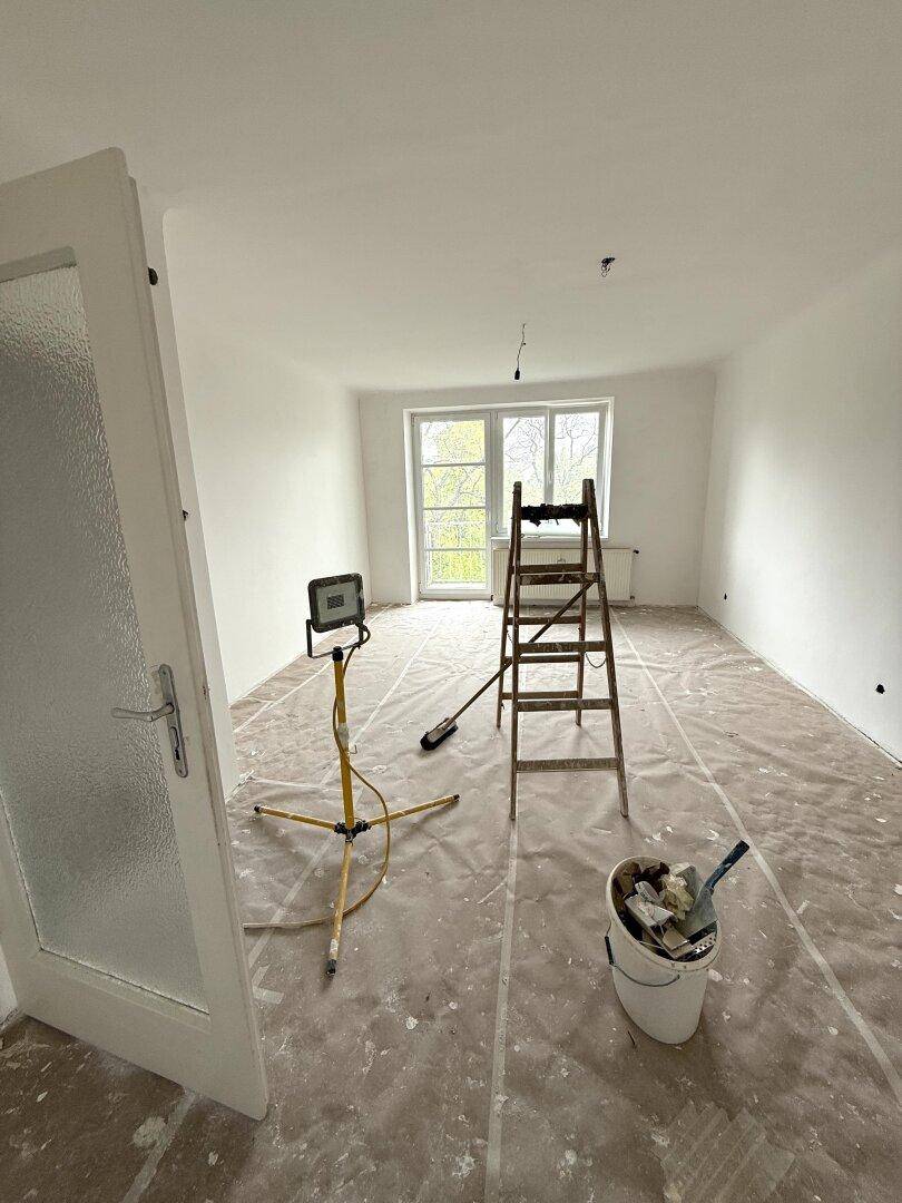 Heller Raum in Renovierung mit Zugang zu einem Balkon, weißen Wänden und einer Arbeitslampe auf dem abgedeckten Boden.