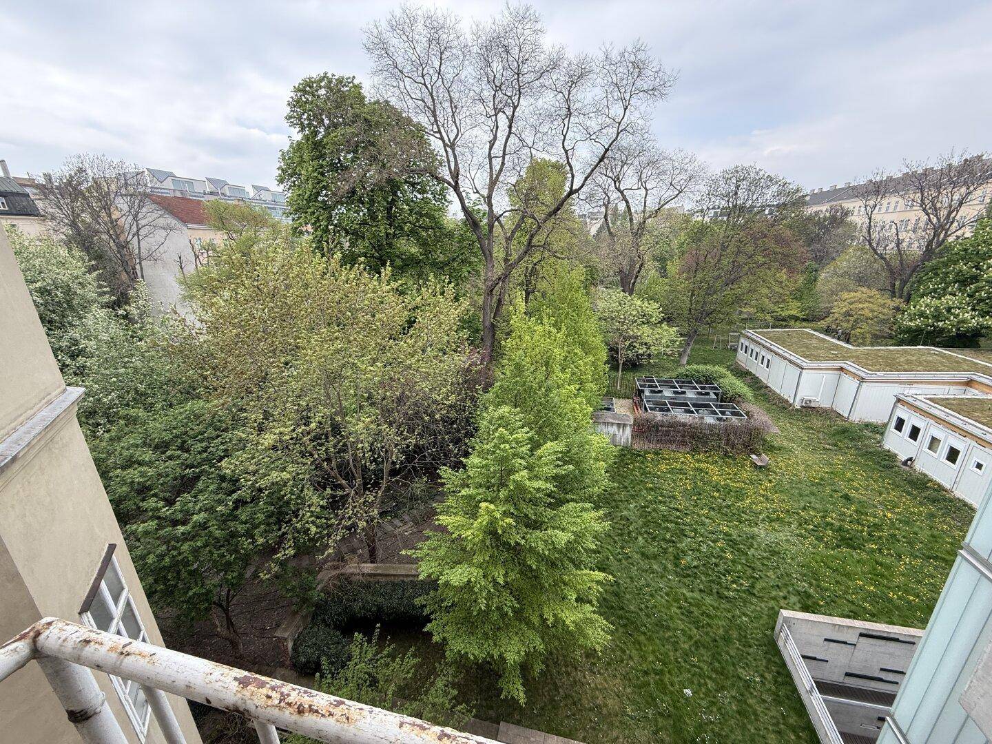 Weitläufiger Blick auf eine grüne Parklandschaft mit altem Baumbestand und niedrigen Gebäuden.