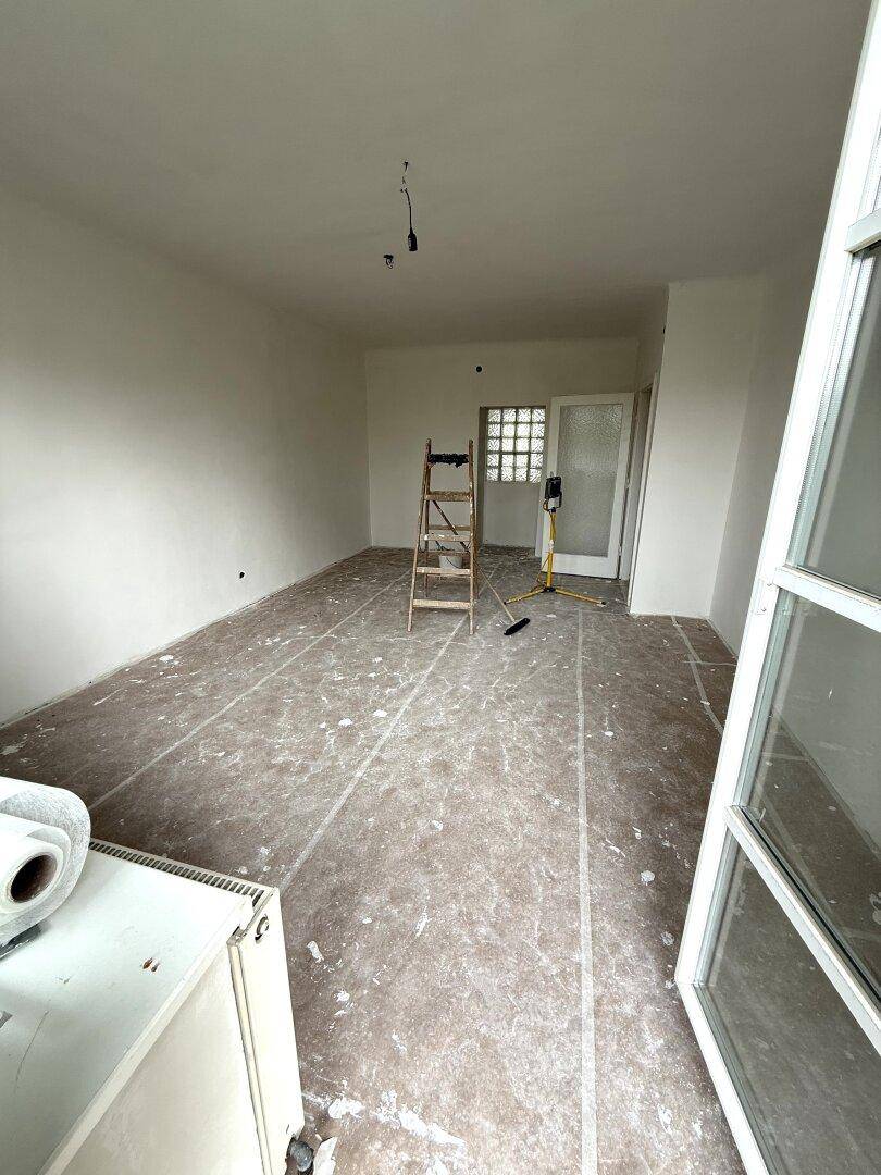 Renovierungsraum mit weißen Wänden, einer Leiter und einem Glasbausteinfenster, das natürliches Licht hereinlässt.