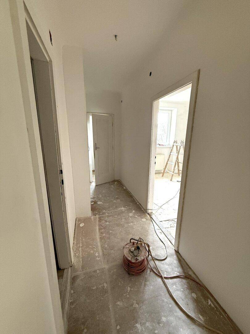 Renovierungsbedürftiger Flur mit weißen Wänden, offenen Türrahmen und einem Kabel auf dem abgedeckten Boden.