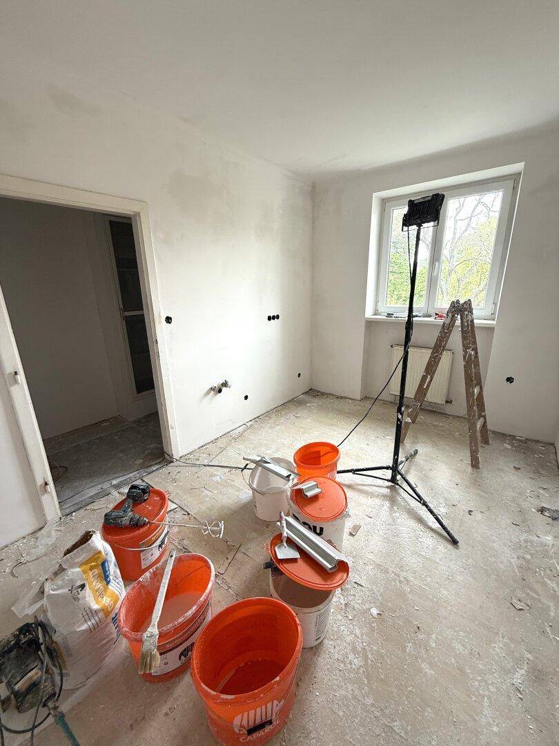 Renovierungsraum mit weißen Wänden, einem Fenster, Arbeitsbeleuchtung und Baumaterialien auf dem abgedeckten Boden.
