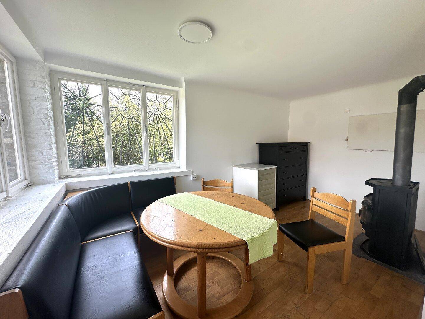 Gemütlicher Wohnbereich mit Holzofen, rundem Tisch und Erkerfenster mit Blick ins Grüne.