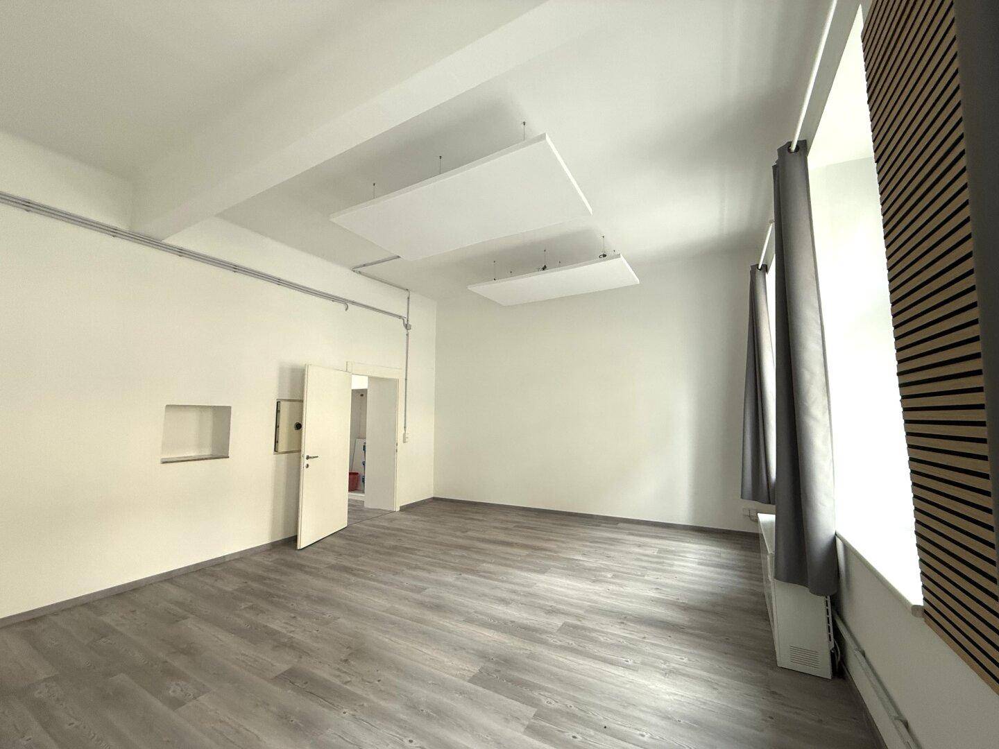 Heller, leerer Raum mit Laminatboden, weißen Wänden und schwebenden Akustikpaneelen an der Decke.