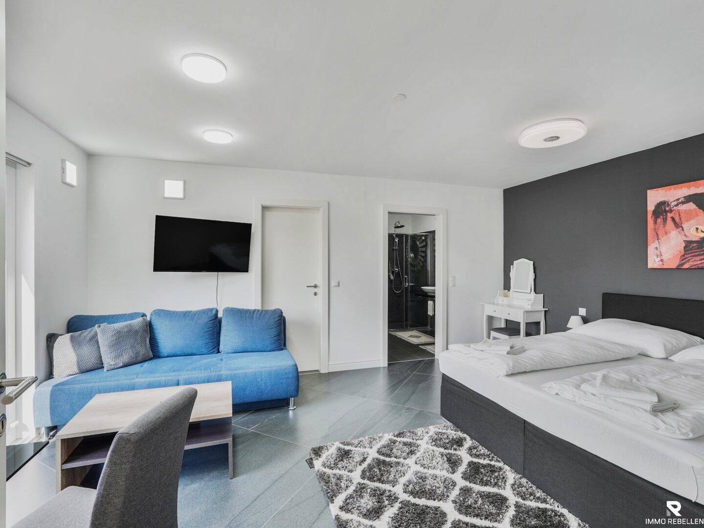Helles Schlafzimmer mit Sofa, Fernseher und Zugang zu einem weiteren Badezimmer.