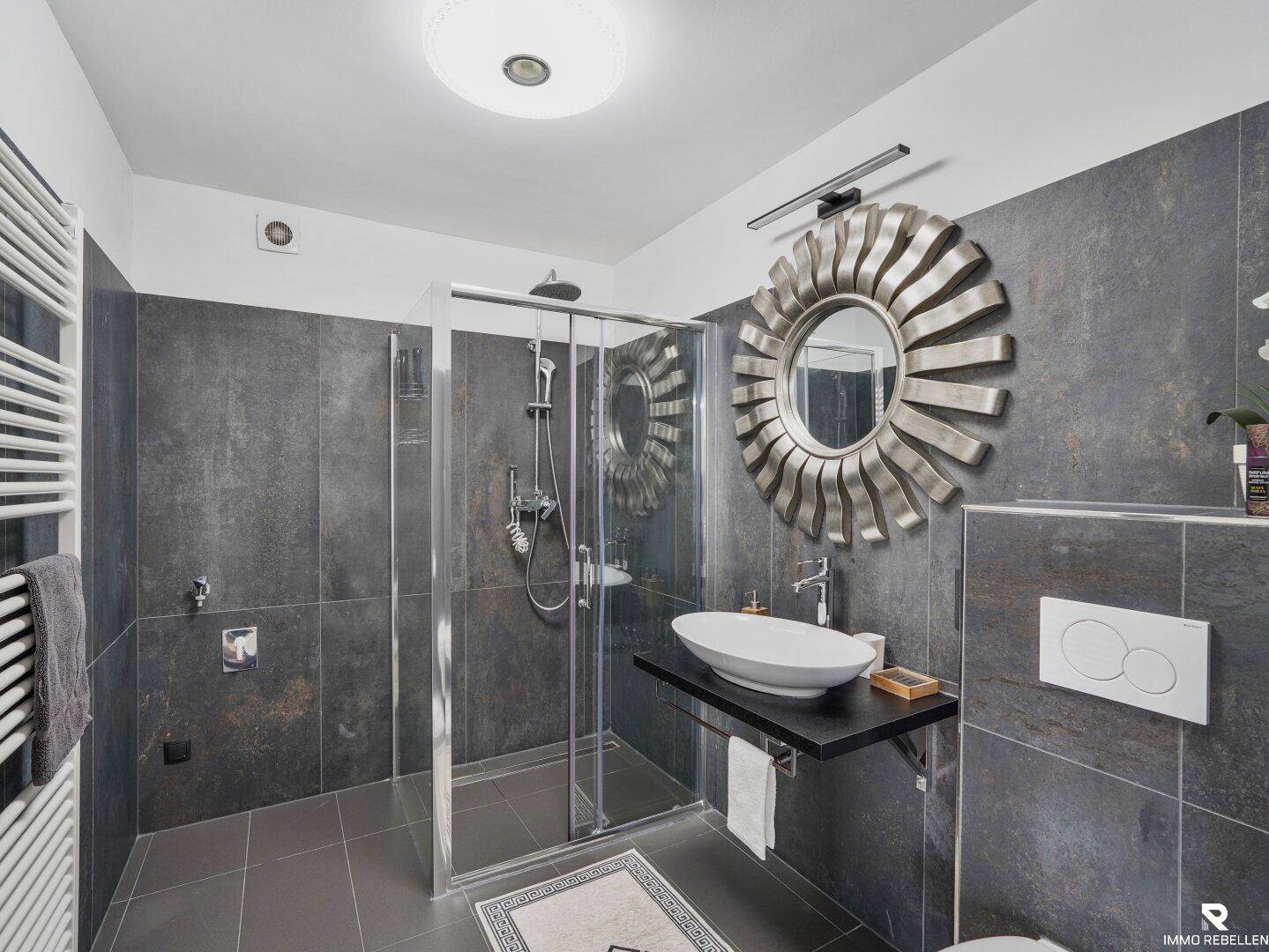 Modernes Badezimmer mit ebenerdiger Dusche, beheiztem Handtuchhalter und dunklen Fliesen.