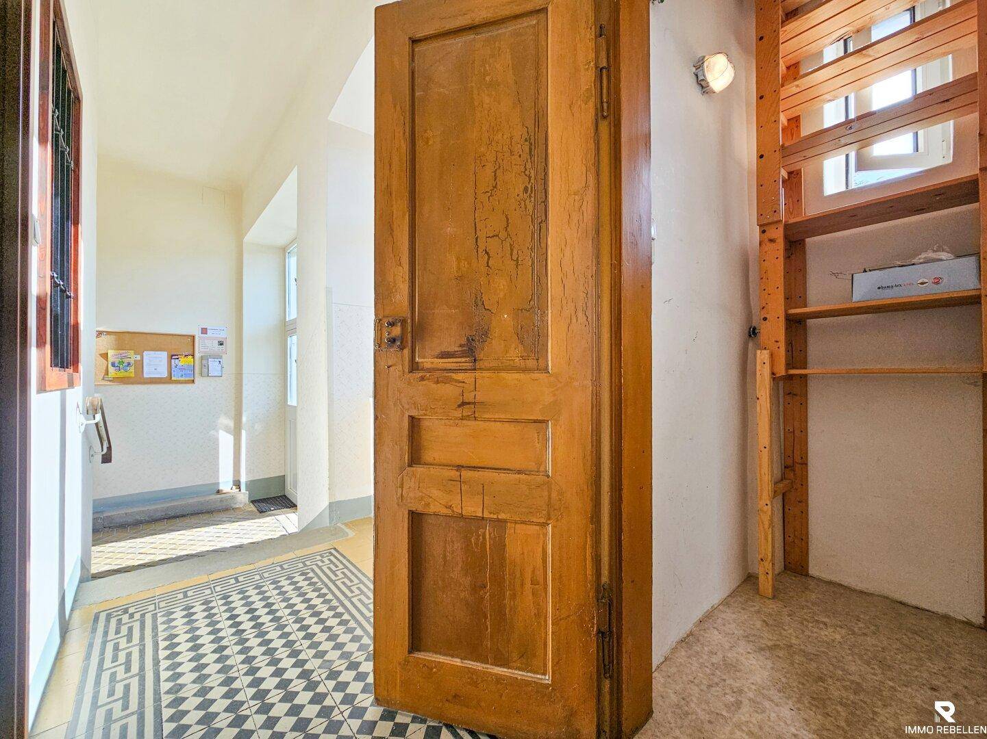 Heller Treppenhausflur mit gemusterten Fliesen und einer alten, massiven Holztür.