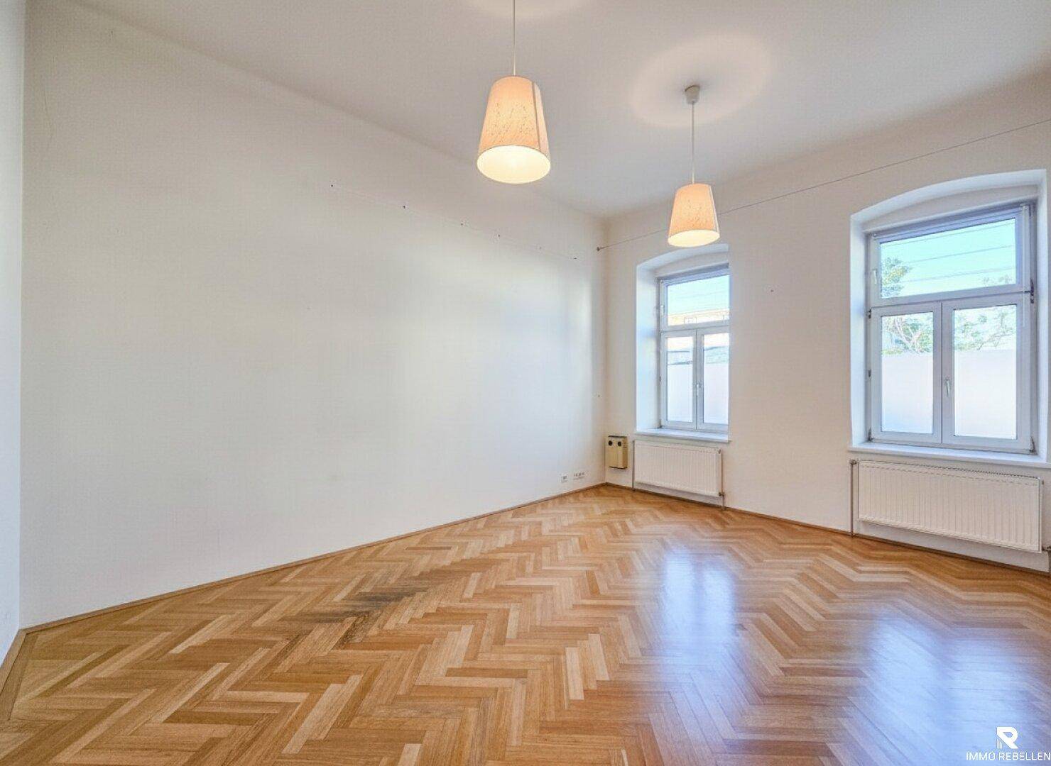 Helles, leeres Zimmer mit klassischem Fischgrätparkettboden und zwei großen Fenstern.