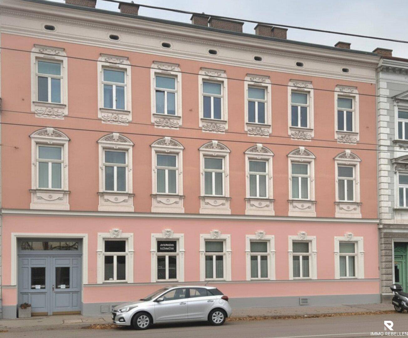 Mehrstöckiges Wohngebäude mit rosa Fassade, verzierten Fenstern und einem geparkten Auto davor.