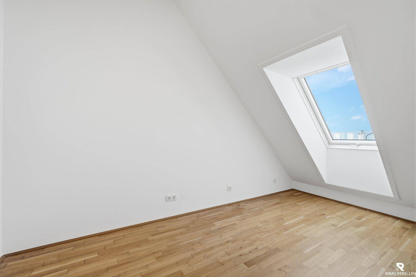 Leeres Schlafzimmer mit Holzboden, weiß gestrichenen Wänden und einem großen Dachfenster.