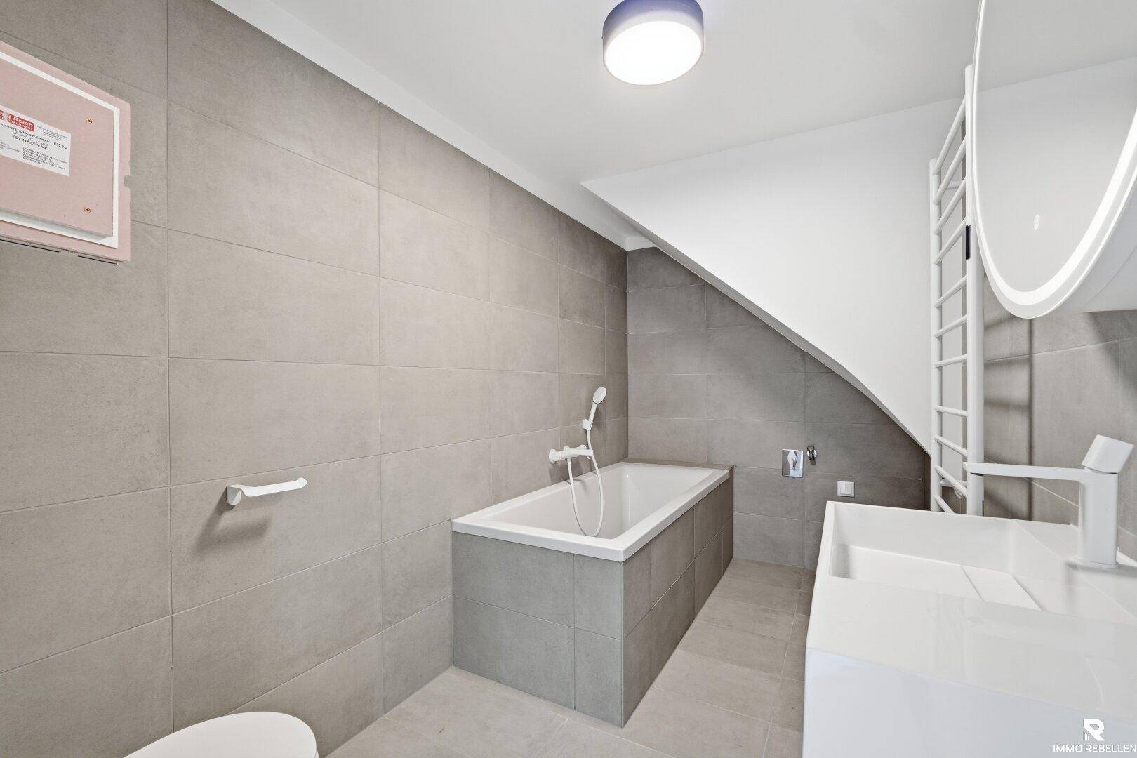 Modernes Badezimmer mit Badewanne, grauen Fliesen, Waschbecken und beheiztem Handtuchhalter.