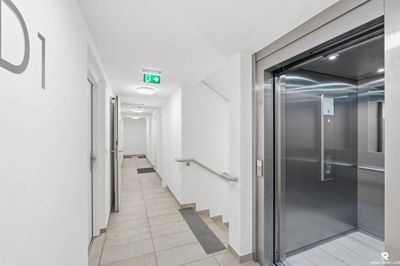 Heller Flur im Gebäude mit Fliesenboden, Handlauf und einem modernen Aufzug.